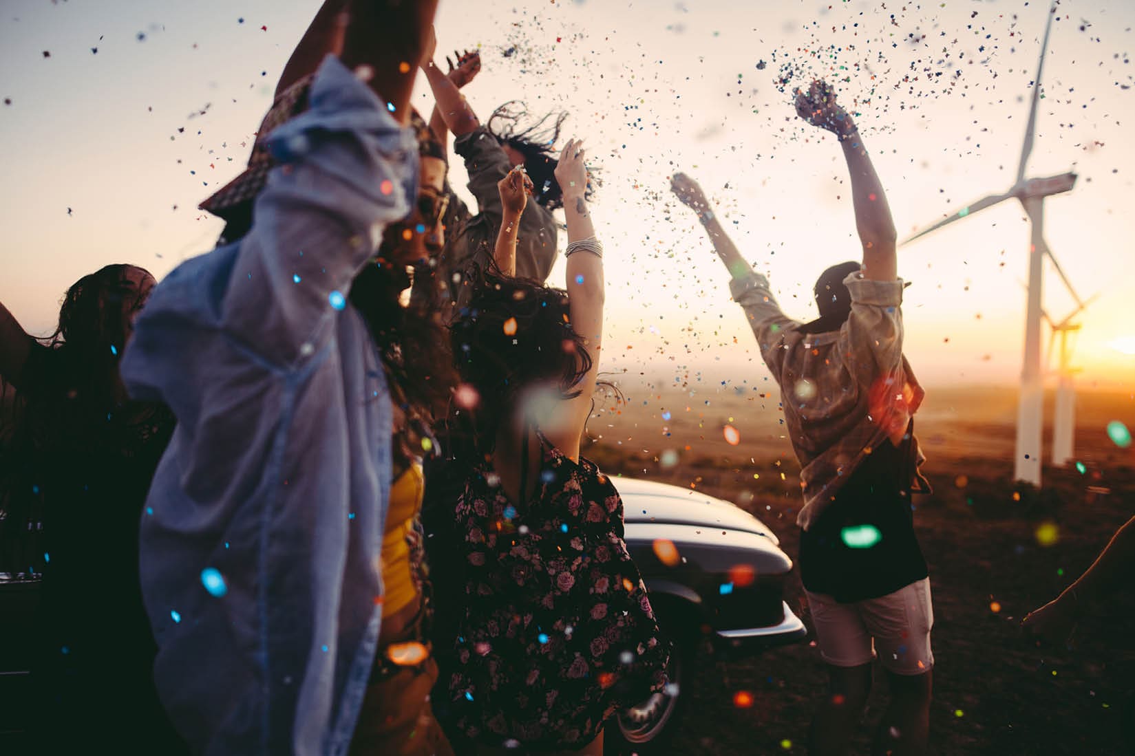 Group of multi-ethnic teens dressed in boho style partying outdoors throwing colourful confetti in a wind farm at sunset during their american carefree road trip
